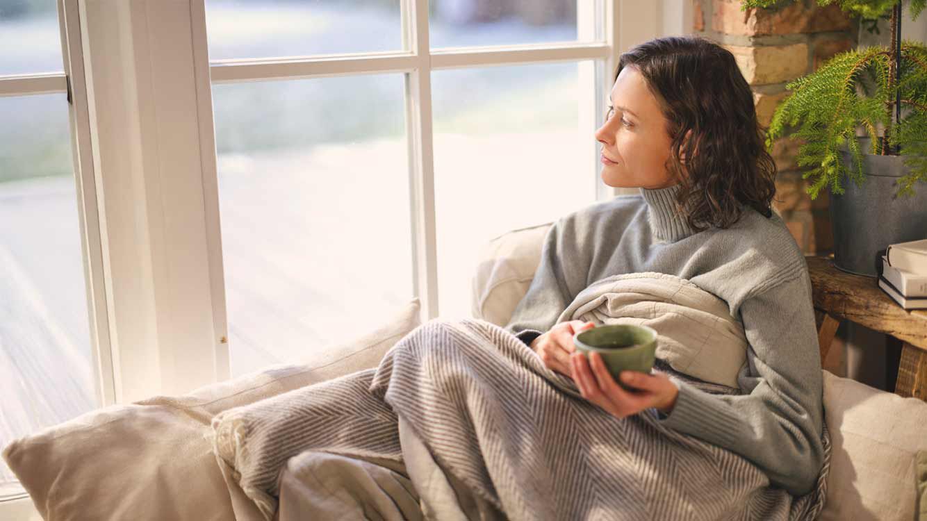 Een vrouw zit met een kopje thee, lekker onder een deken gekruipt, bij het raam. 